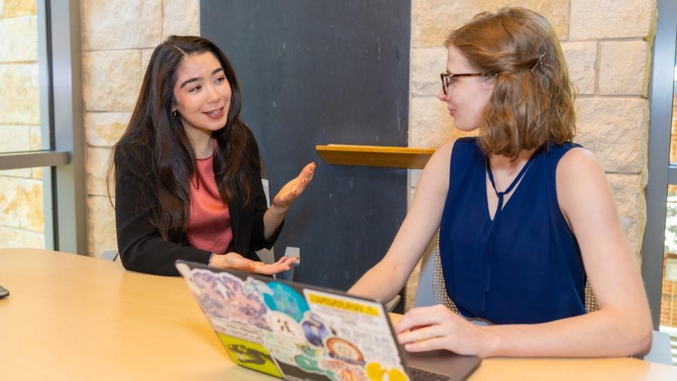 Two students talk together at a wooden table with a laptop